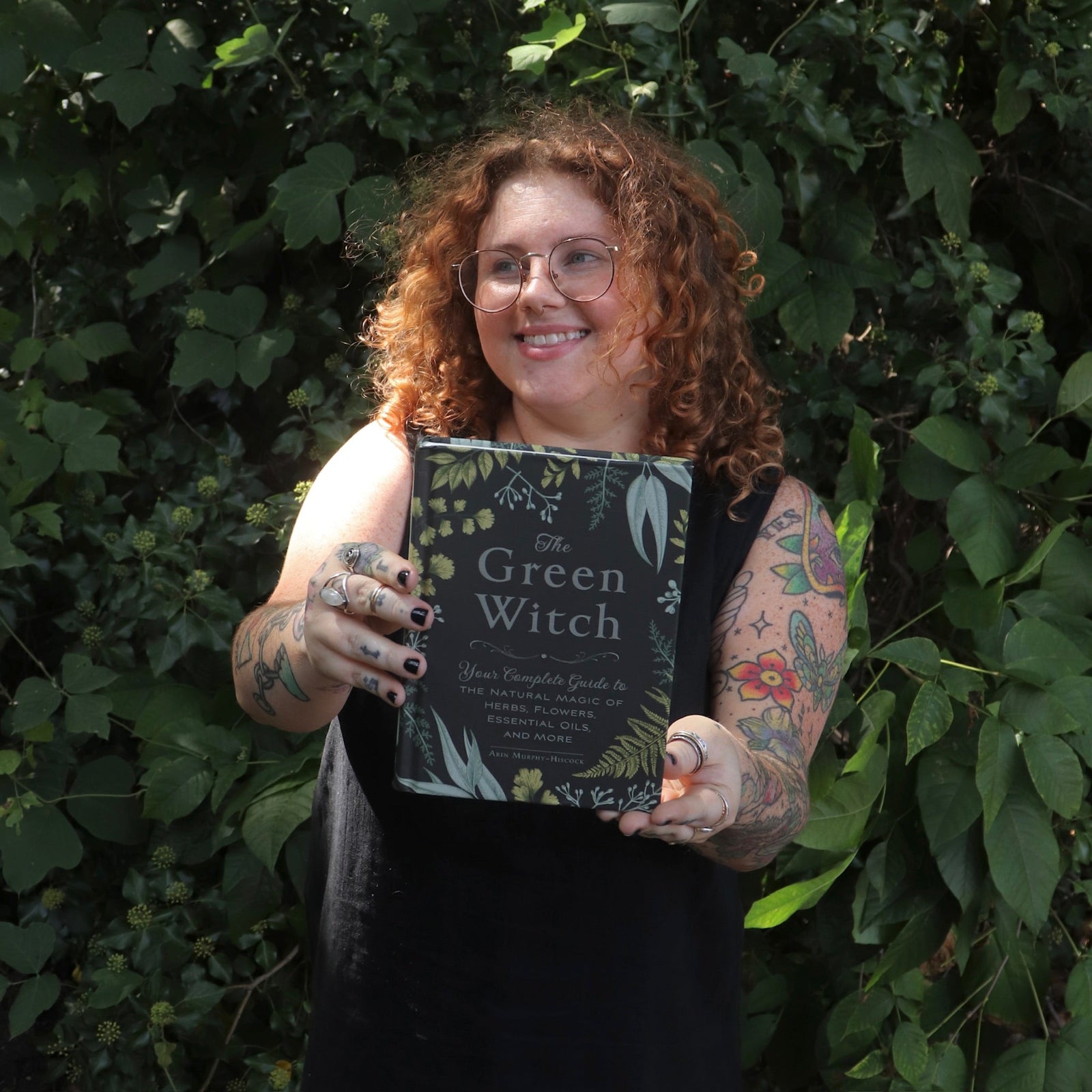 A woman holding the Green Witch by Arin Murphy-Hiscock in front of greenery.