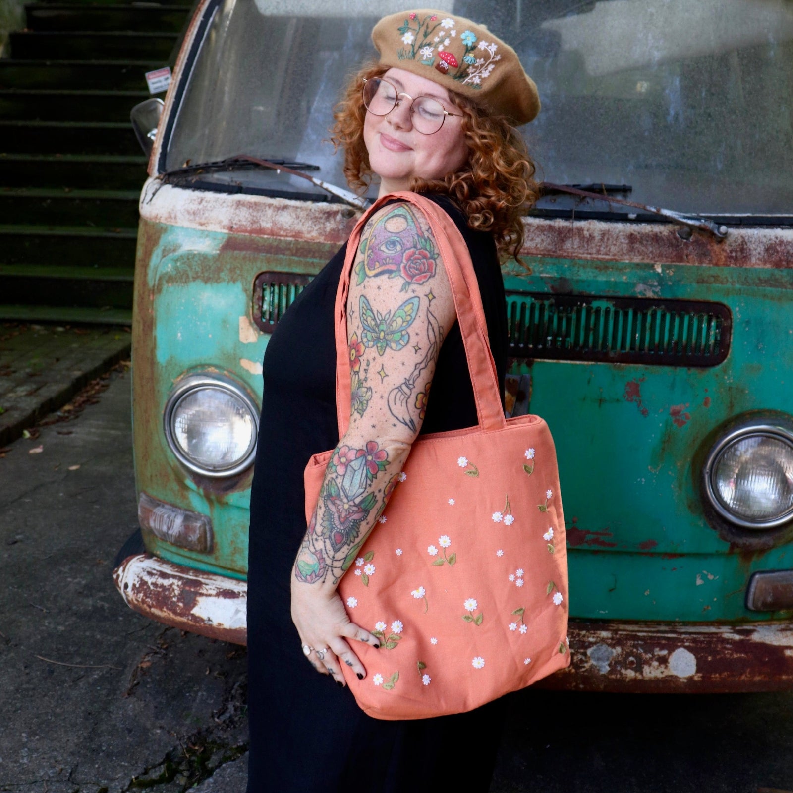 A woman has the Daisy Embroidered Orange Linen Zipper Tote Bag on her shoulder while wearing a black tank dress.