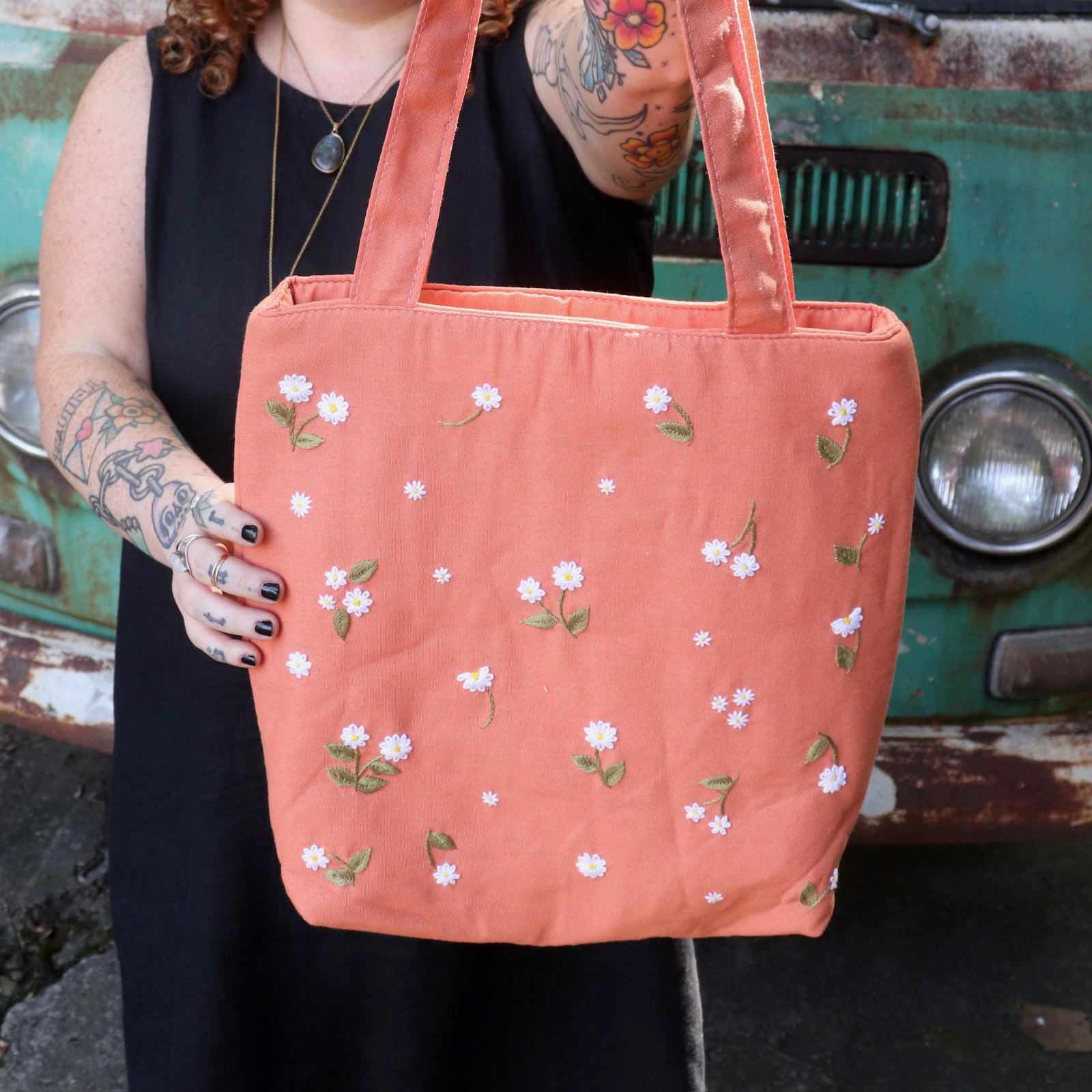 A woman holding the Daisy Embroidered Orange Linen Zipper Tote Bag while wearing a black tank dress.