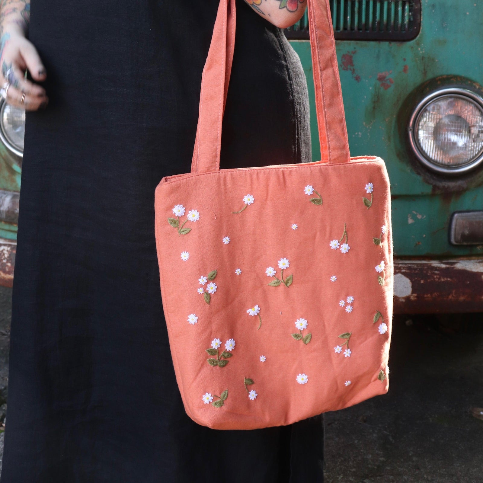 A woman holding the Daisy Embroidered Orange Linen Zipper Tote Bag while wearing a black tank dress.