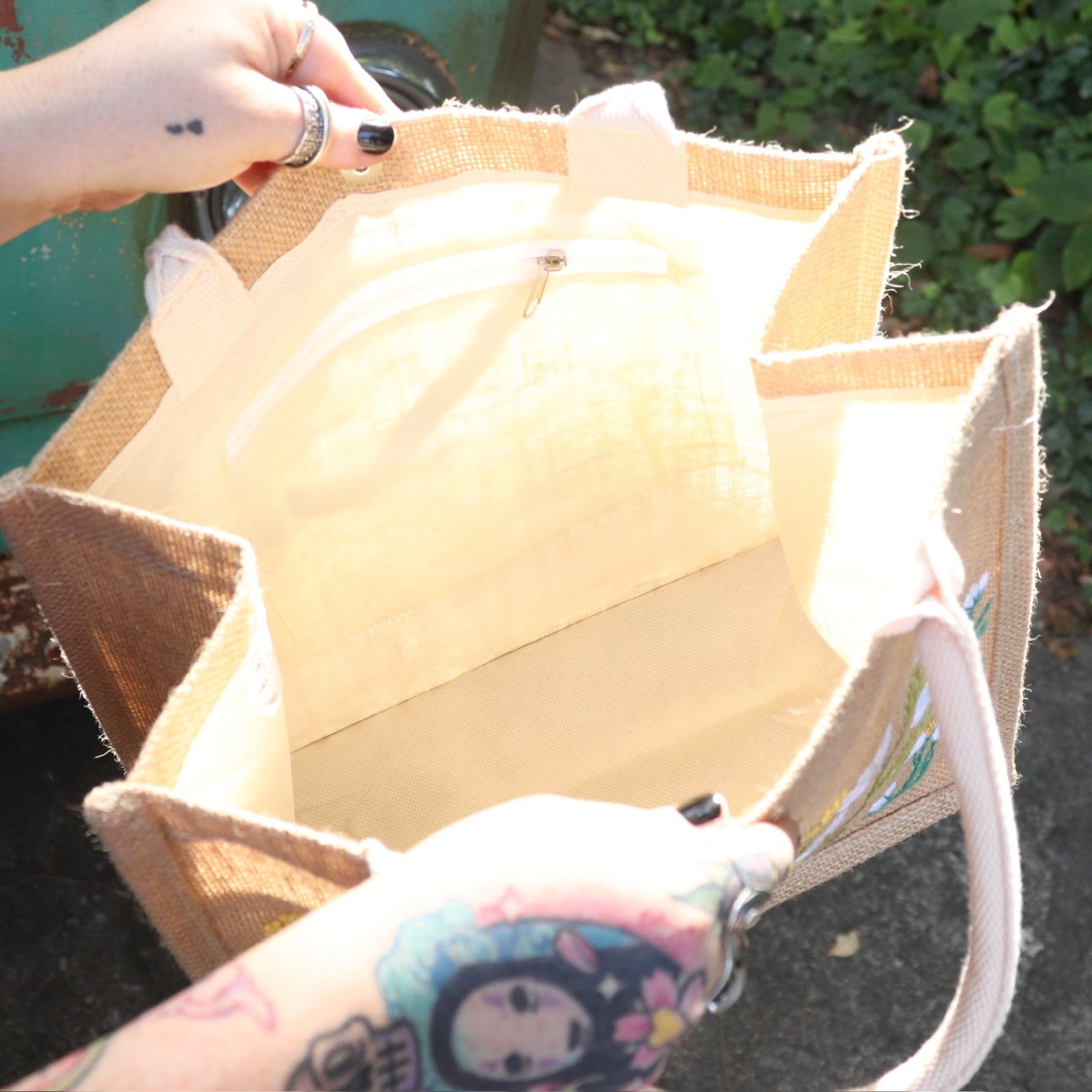 A woman wearing a black tank dress holding the Daisy Garden Embroidered Jute Bag open showing off the inside.