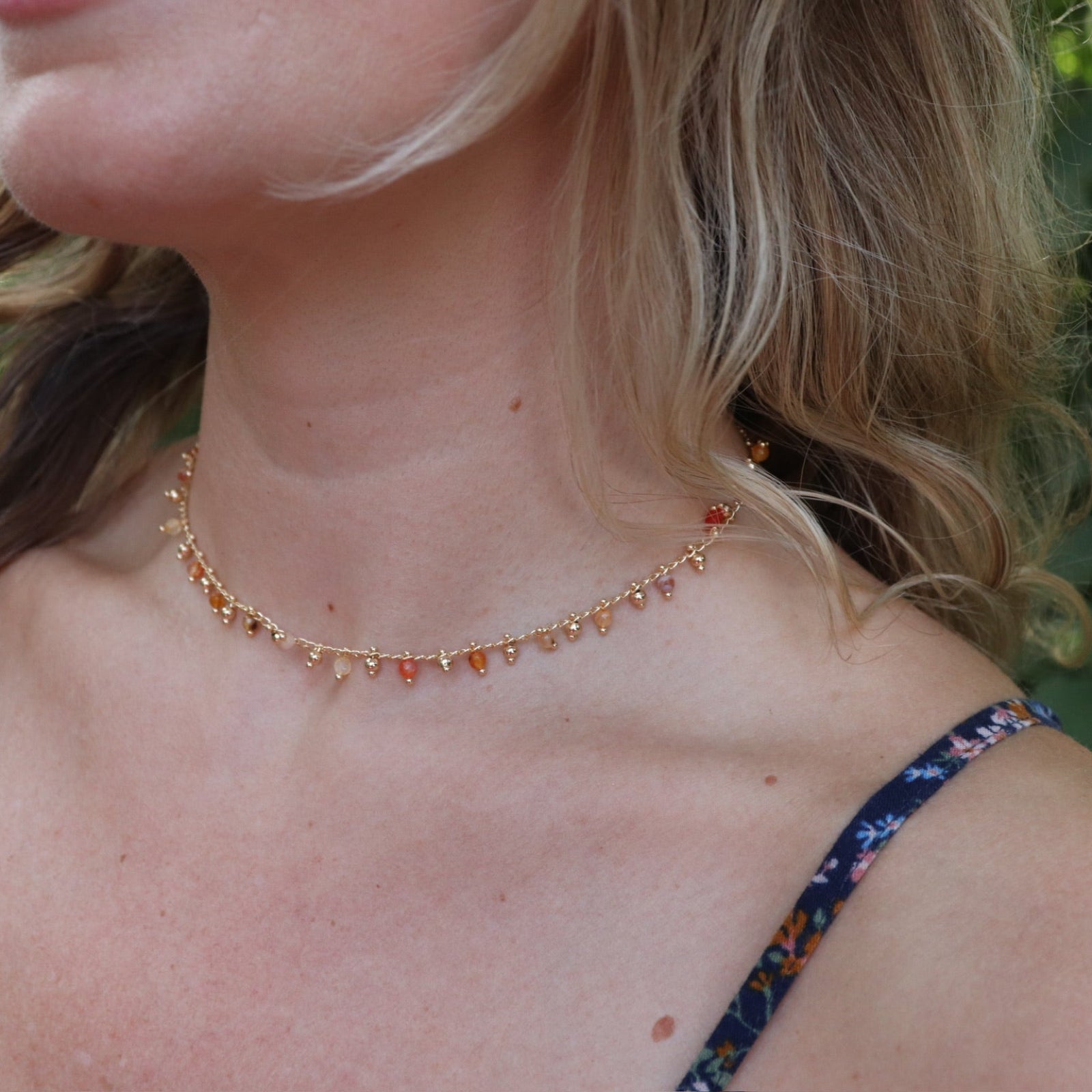 A woman wearing the orange Midsummer Gemstone Agate Gold Plated Necklace in front of some greenery.