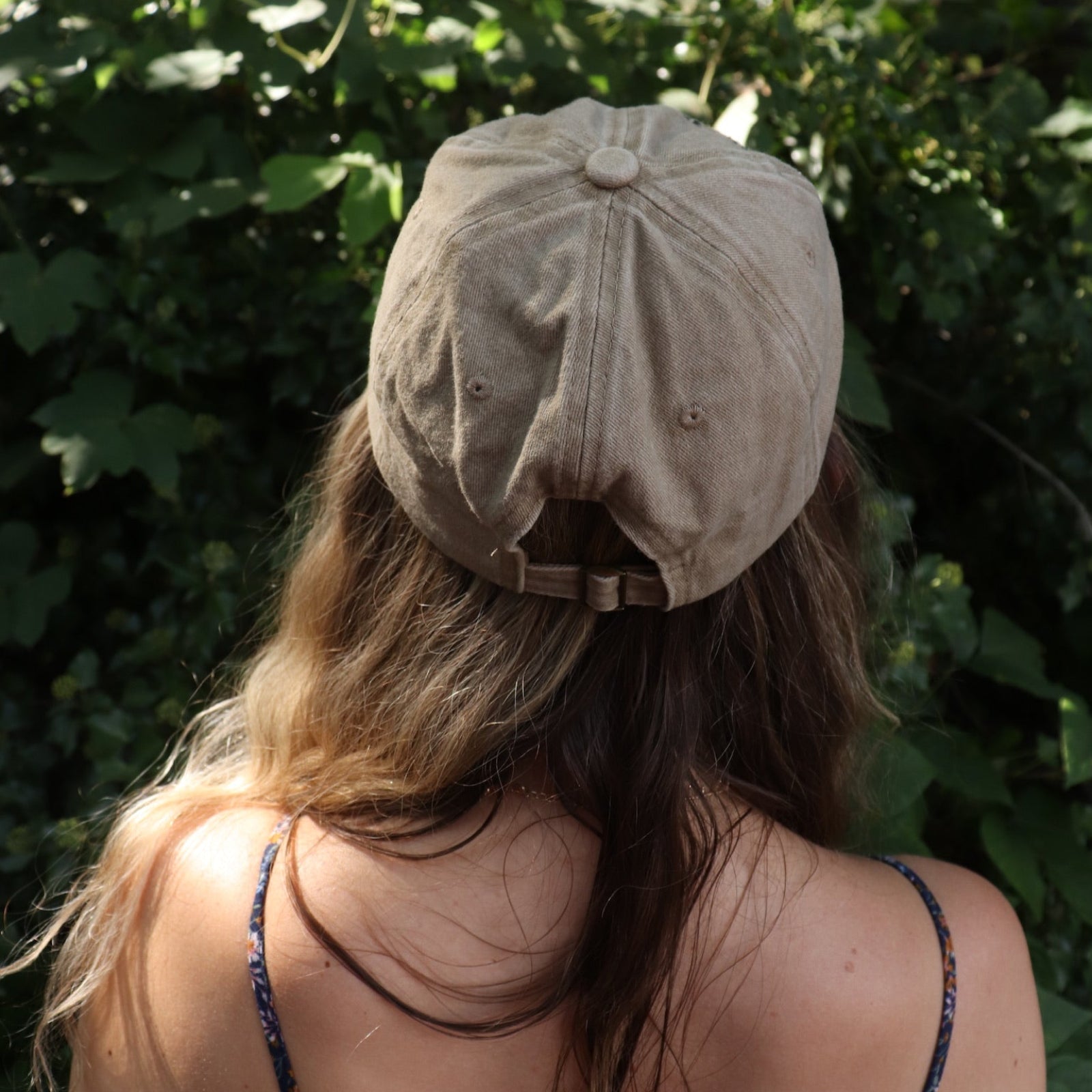 A woman wearing the Mushroom Forest & Bee Embroidered Adjustable Baseball Cap with a navy floral dress in front of greenery, showing the back.