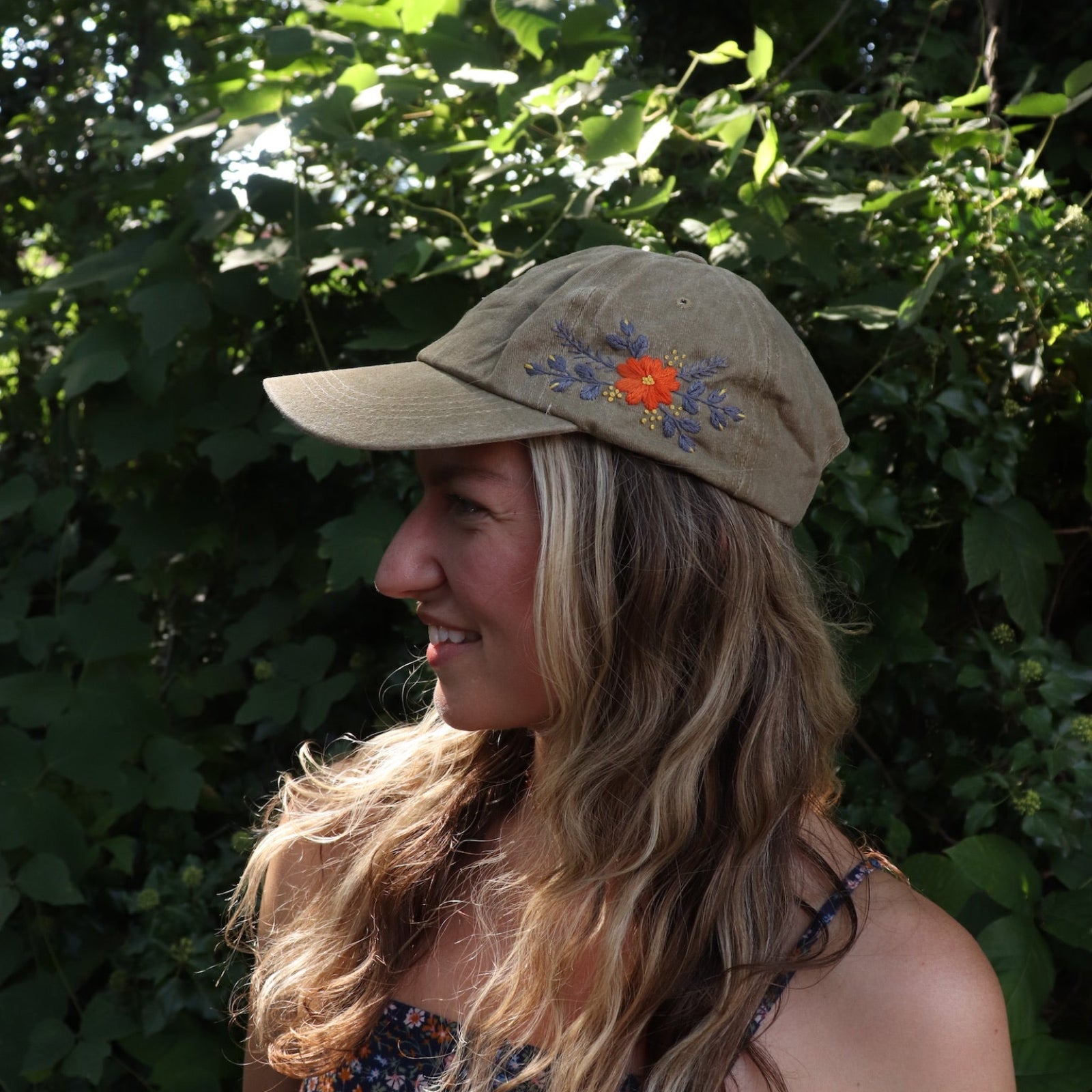 A woman wearing the Orange Pansy Embroidered Adjustable Baseball Cap with a navy floral dress in front of the greenery.
