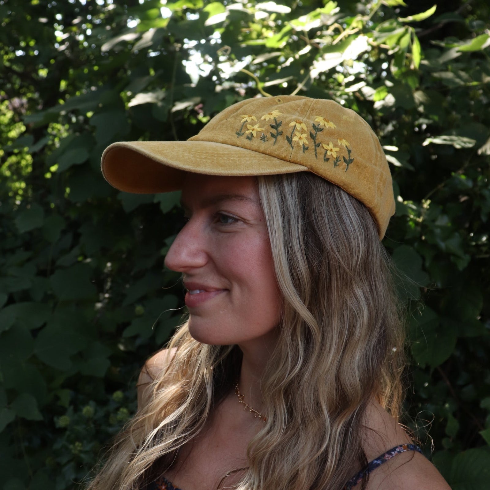 A woman wearing the Daisy Garden Embroidered Adjustable Baseball Cap with a navy floral dress in front of greenery.