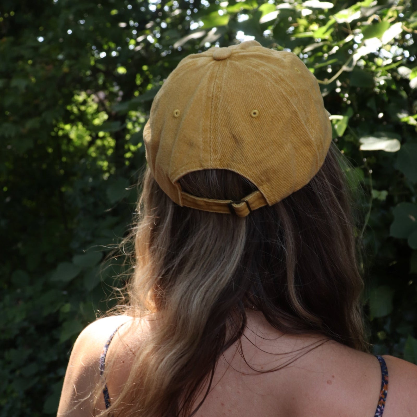 A woman wearing the Daisy Garden Embroidered Adjustable Baseball Cap with a navy floral dress in front of greenery, showing off the back.