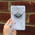 Someone holding the Dainty 4mm Faceted Black Tourmaline Cord 16"-18" Necklace on its card in front of a brick wall.