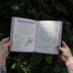 Someone holding the Book of Faerie Spells by Cheralyn Darcey open in front of greenery, showing off the pages.