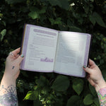 Someone holding the Book of Faerie Spells by Cheralyn Darcey open in front of greenery, showing off the pages.