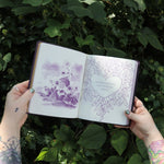 Someone holding the Book of Faerie Spells by Cheralyn Darcey open in front of greenery, showing off the pages.