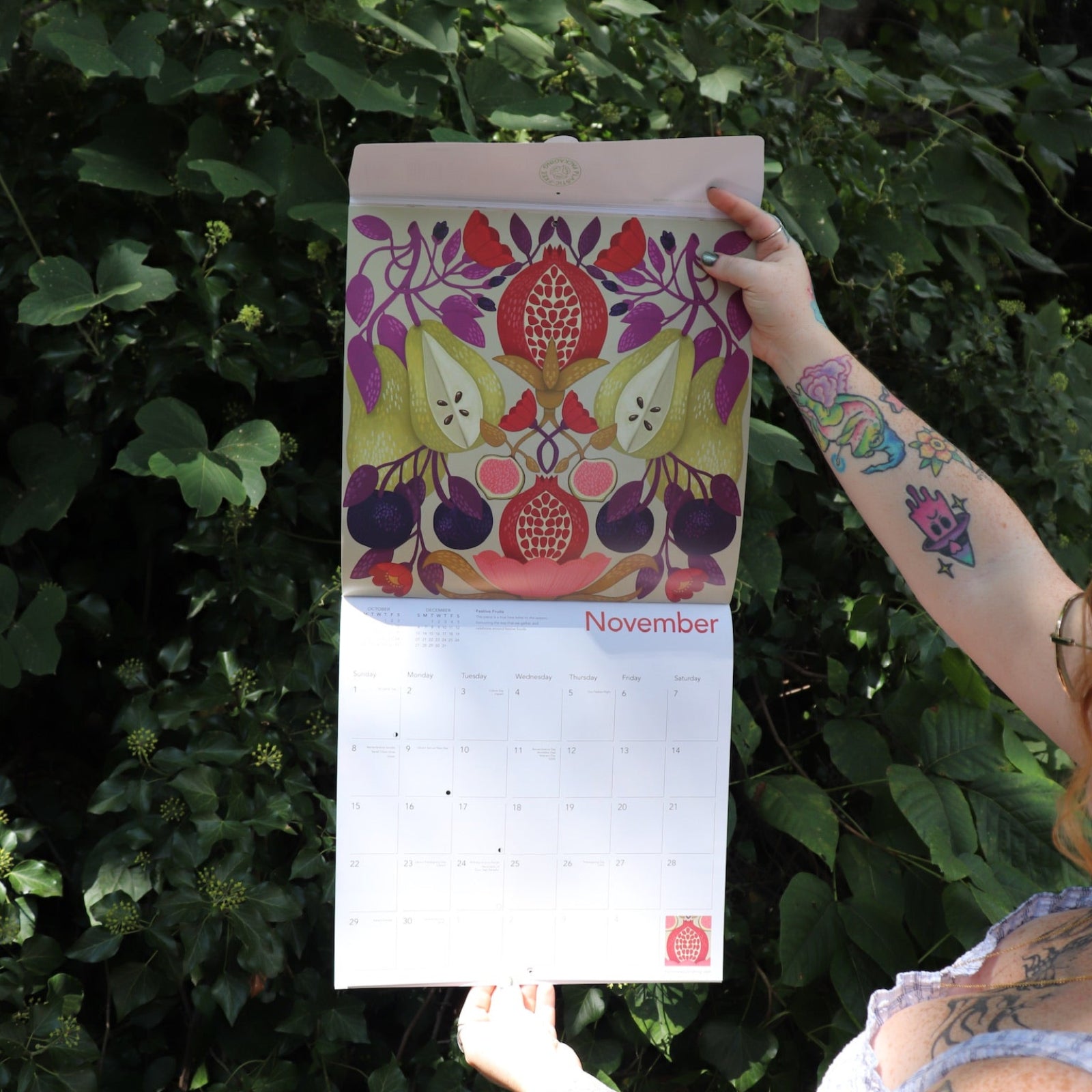 Someone holding the Nina Pace Wall Calendar 2026 open in front of greenery, showing off the pages.