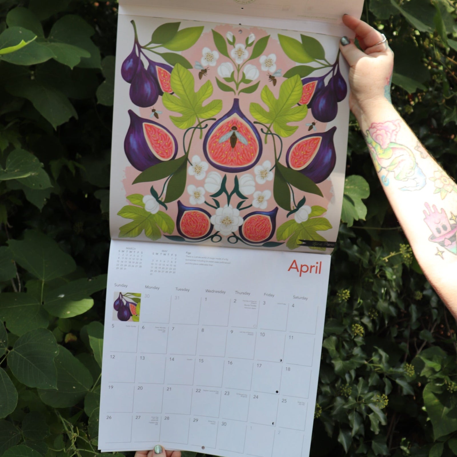 Someone holding the Nina Pace Wall Calendar 2026 open in front of greenery, showing off the pages.