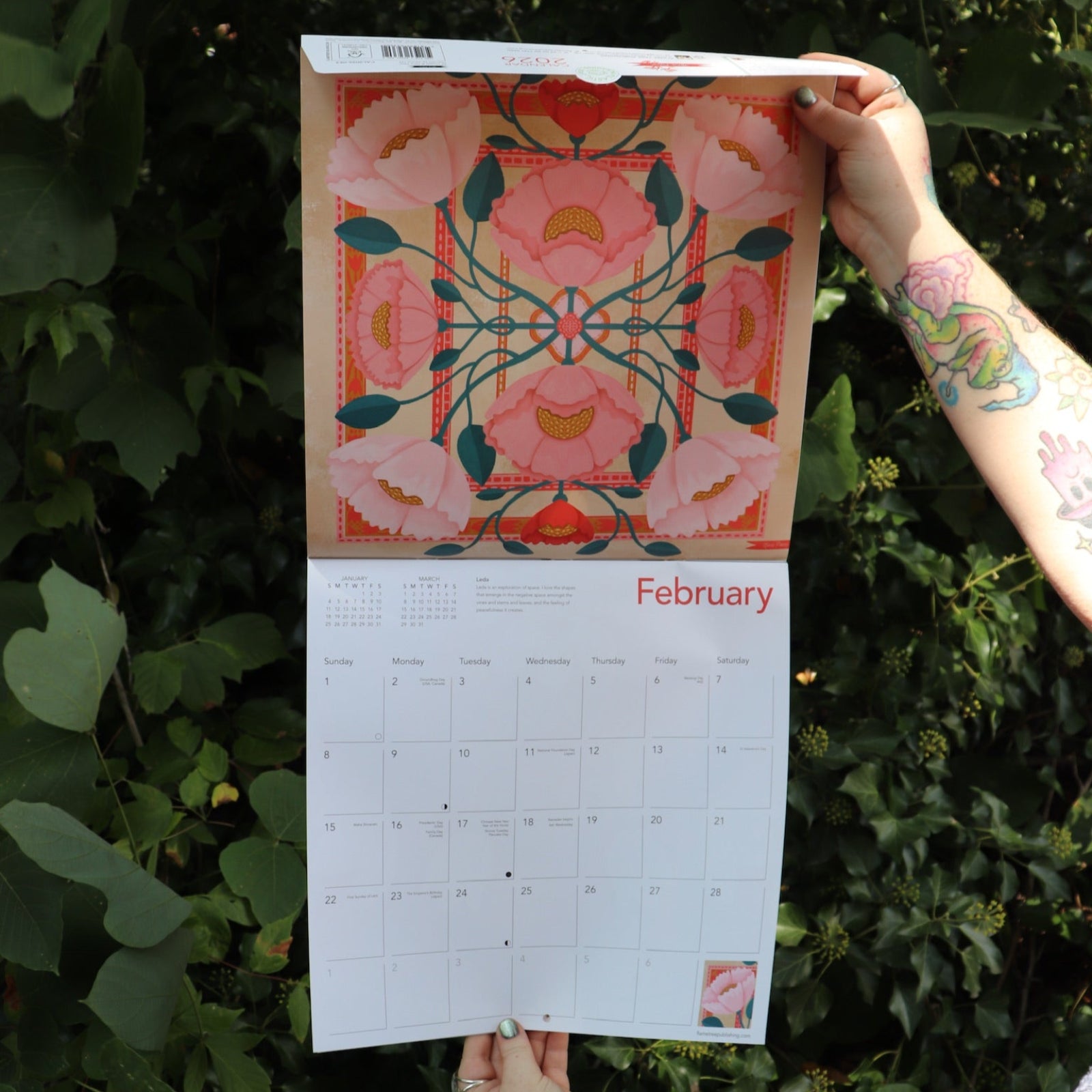 Someone holding the Nina Pace Wall Calendar 2026 open in front of greenery, showing off the pages.