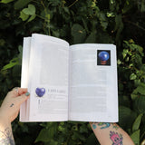 Someone holding the Book of Stones by Robert Simmons open in front of greenery, showing off the pages.
