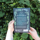 Someone holding the Green Witch's Grimoire by Arin Murphy-Hiscock in front of greenery, showing off the back.