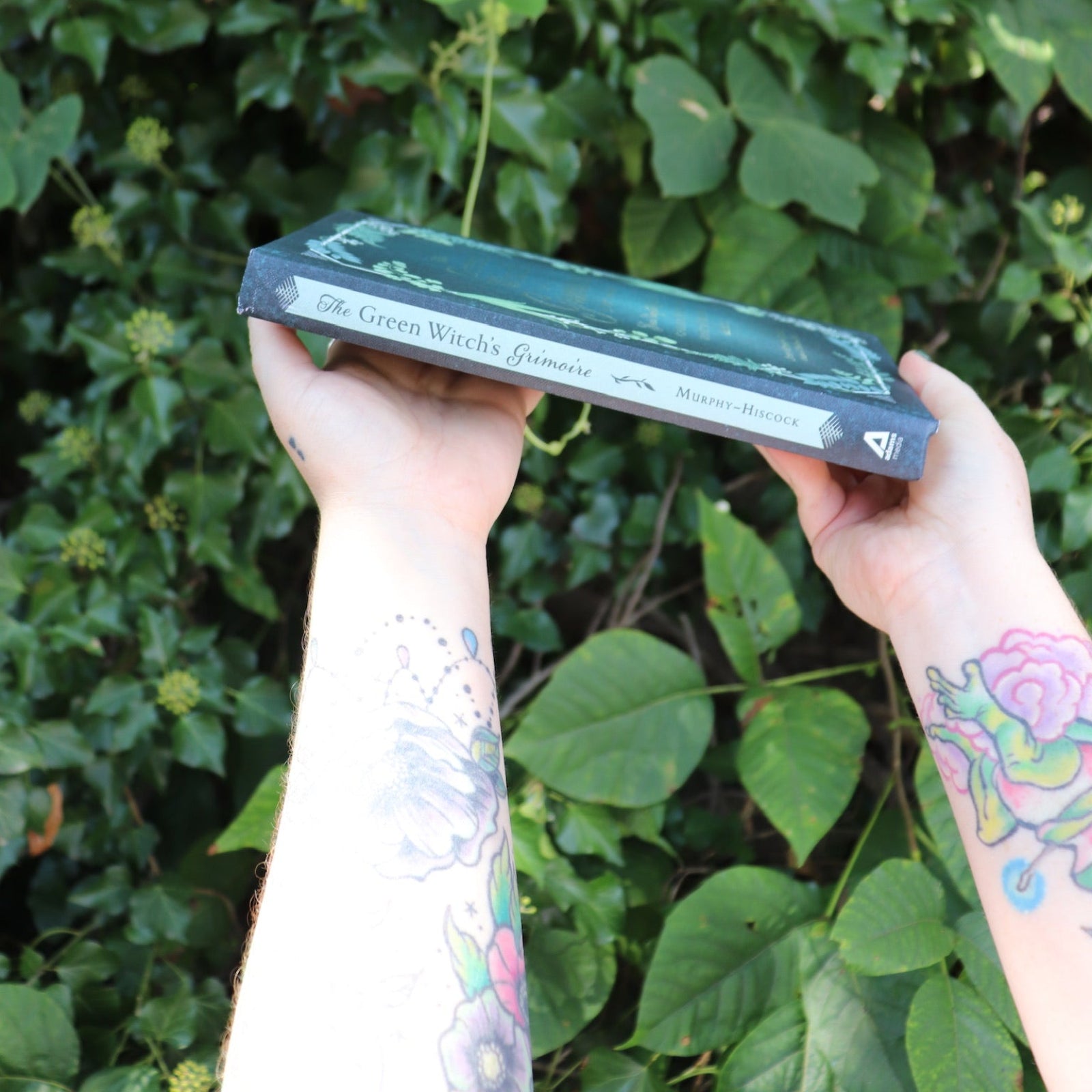 Someone holding the Green Witch's Grimoire by Arin Murphy-Hiscock in front of greenery, showing off the side.