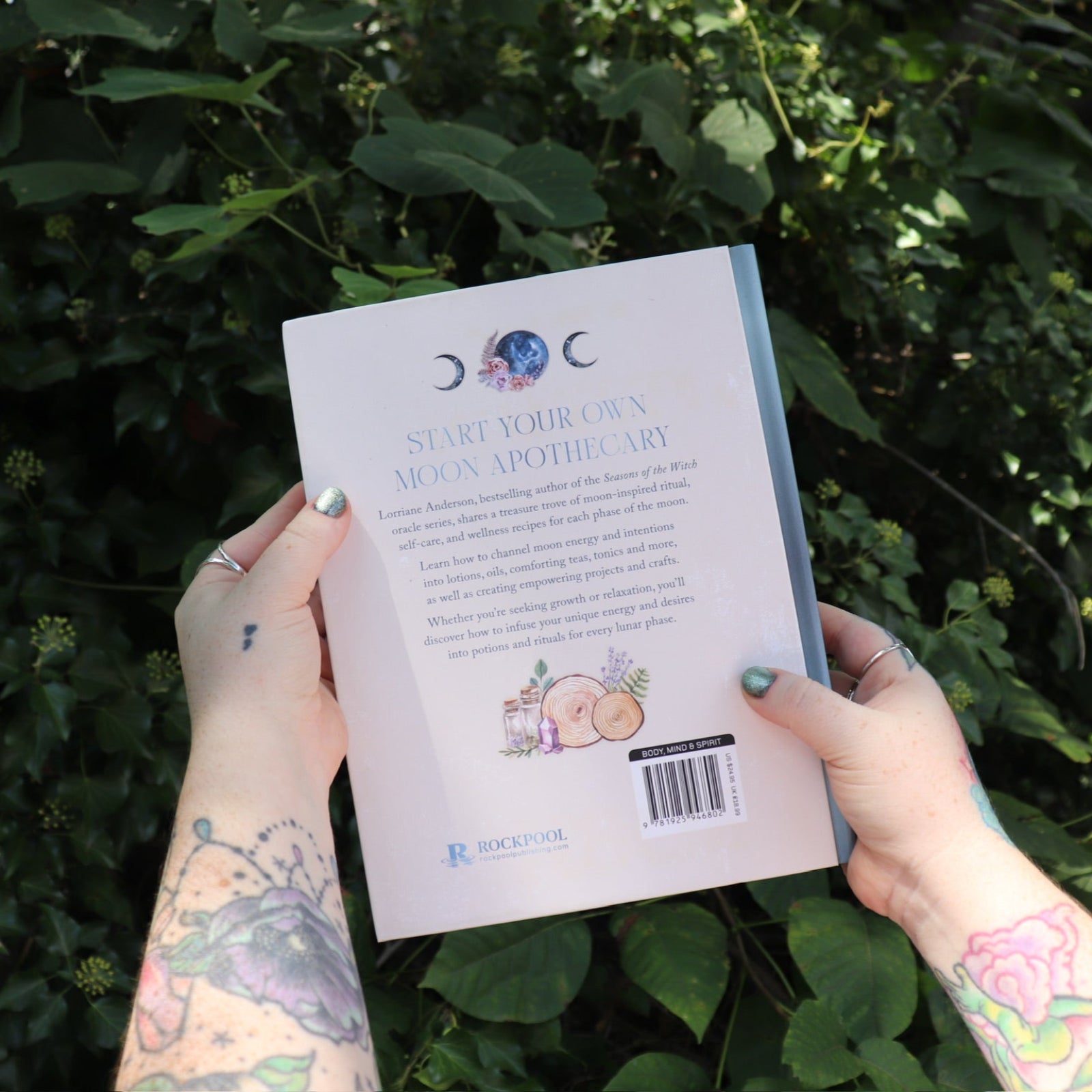 Someone holding the Moon Apothecary by Lorriane Anderson: Hardcover in front of greenery, showing off the back.