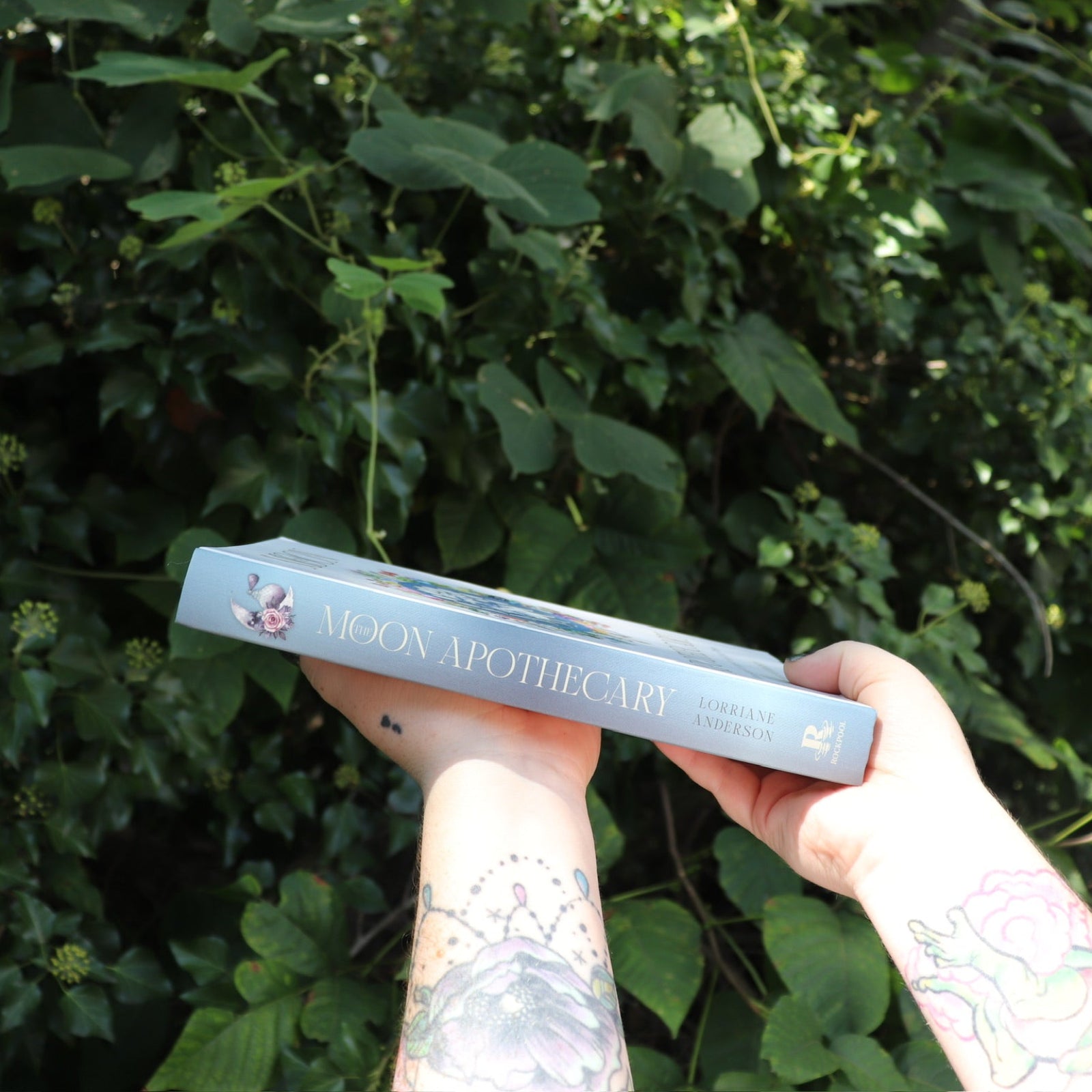 Someone holding the Moon Apothecary by Lorriane Anderson: Hardcover in front of greenery, showing off the side.