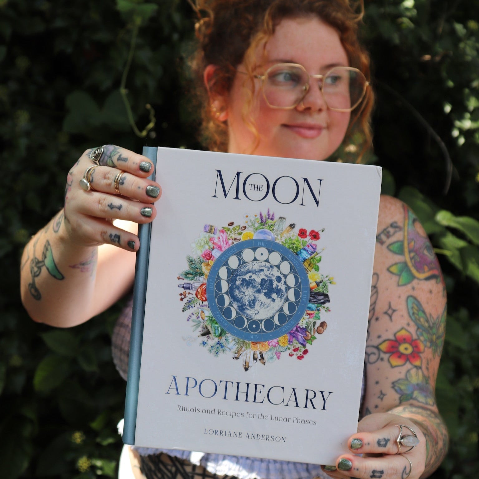 A woman holding the Moon Apothecary by Lorriane Anderson: Hardcover in front of greenery.