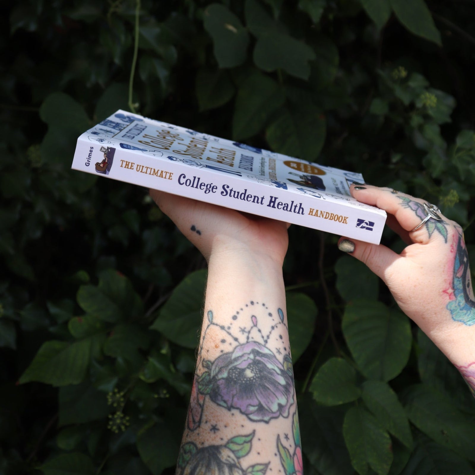 Someone holding the Ultimate College Student Health Handbook by Jill Grimes in front of greenery, showing off the side.