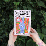 Someone holding the Age of Magical Overthinking Hardcover Book in front of greenery.