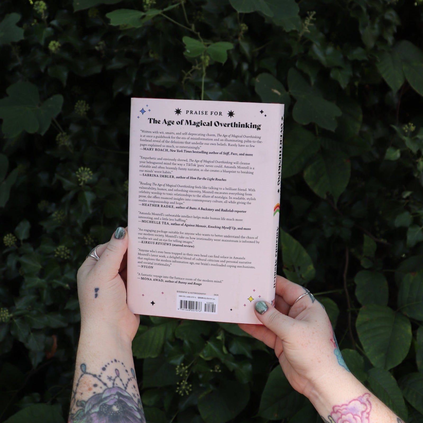 Someone holding the Age of Magical Overthinking Hardcover Book in front of greenery, showing off the back.