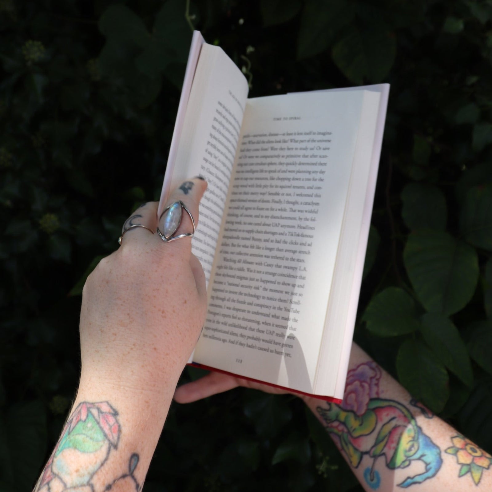 Someone holding the Age of Magical Overthinking Hardcover Book open in front of greenery, showing off the pages.