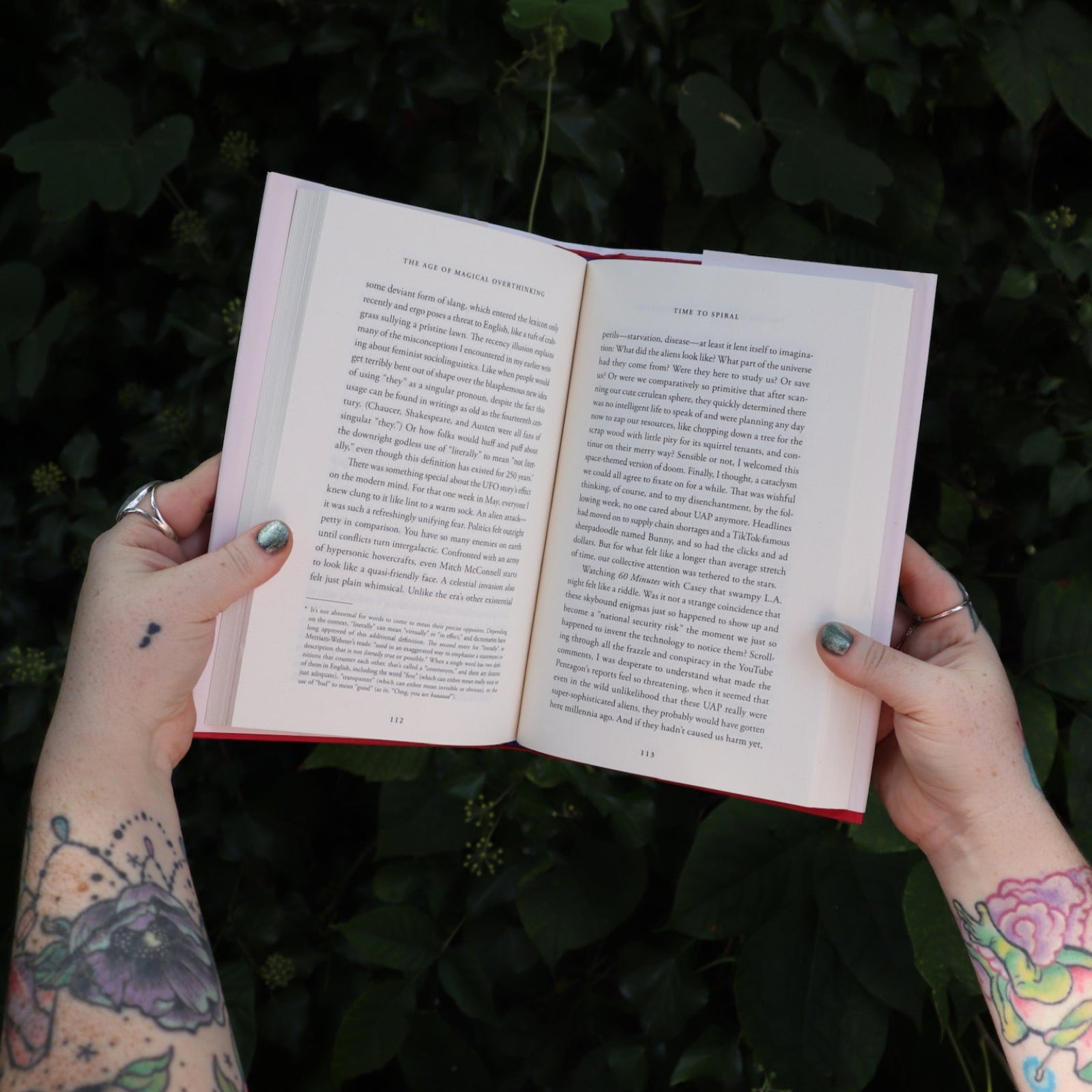 Someone holding the Age of Magical Overthinking Hardcover Book open in front of greenery, showing off the pages.