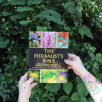 Someone holding the Herbalist's Bible by Julie Bruton-Seal - Paperback Book in front of greenery.