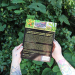 Someone holding the Herbalist's Bible by Julie Bruton-Seal - Paperback Book in front of greenery, showing off the back.