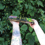 Someone holding the Herbalist's Bible by Julie Bruton-Seal - Paperback Book in front of greenery, showing off the side.