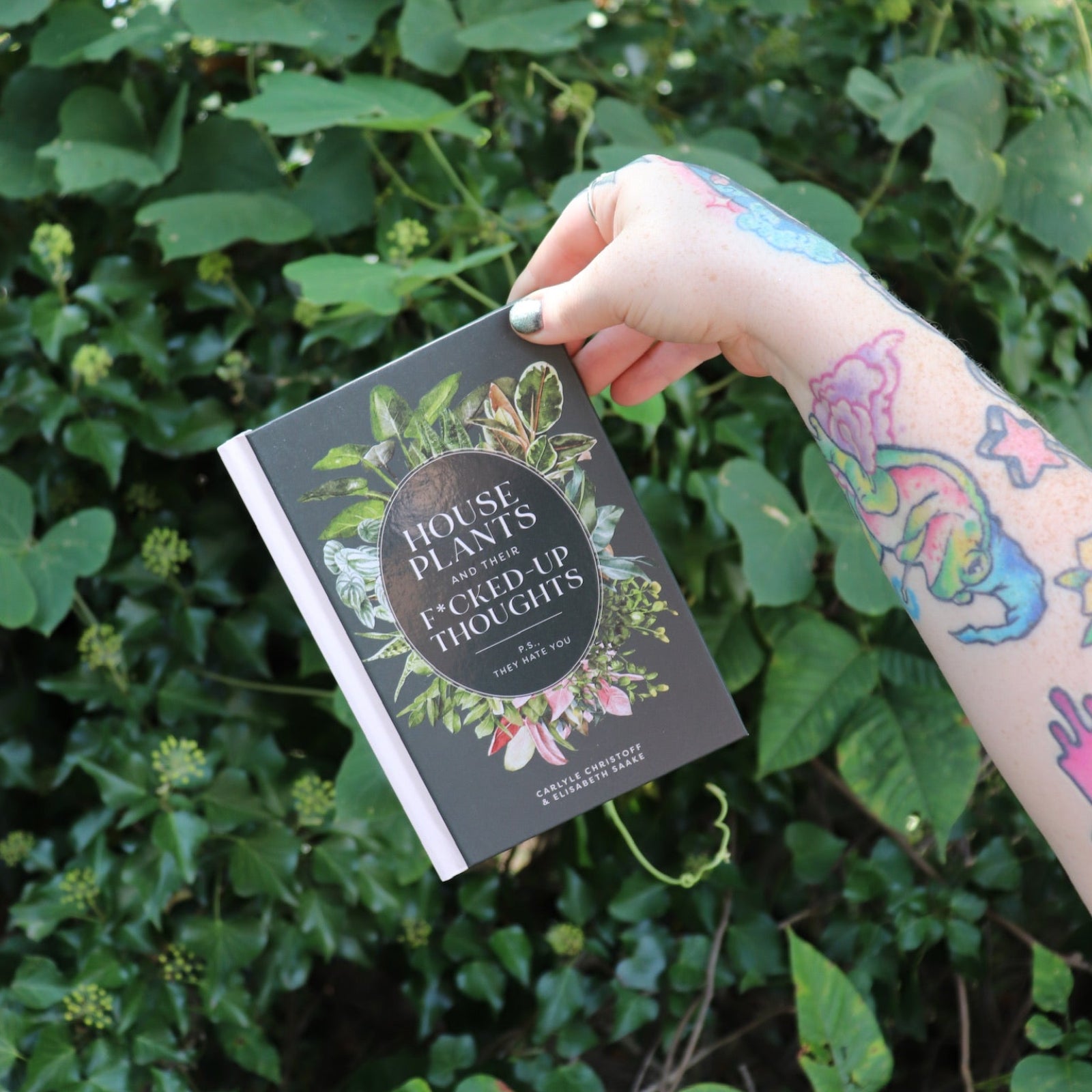 Someone holding the Houseplants and Their F* Up Thoughts by Carlyle Christoff in front of greenery.