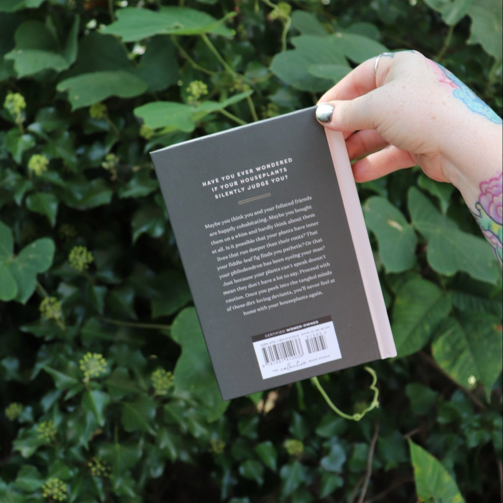 Someone holding the Houseplants and Their F* Up Thoughts by Carlyle Christoff in front of greenery, showing off the back.