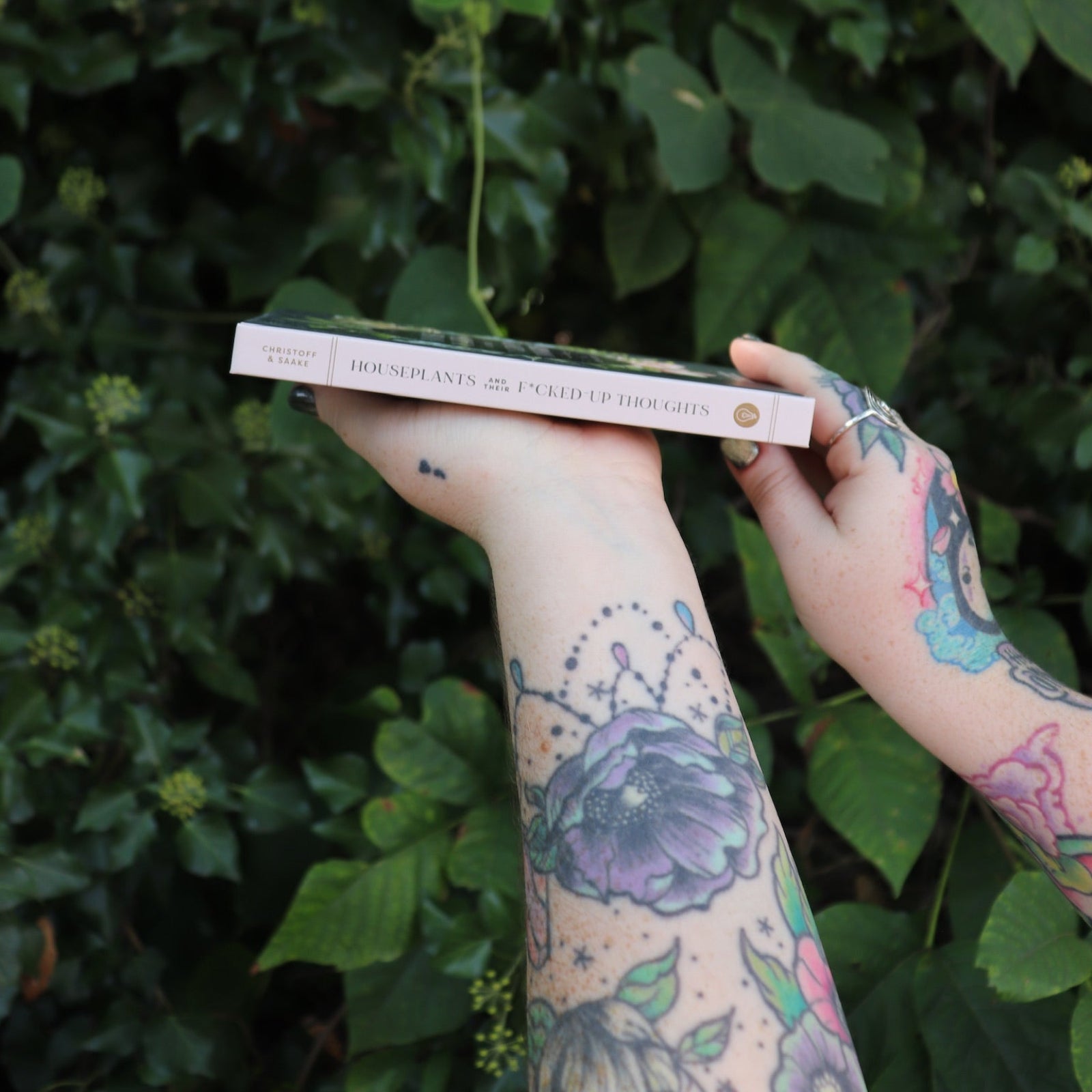 Someone holding the Houseplants and Their F* Up Thoughts by Carlyle Christoff in front of greenery, showing off the side.