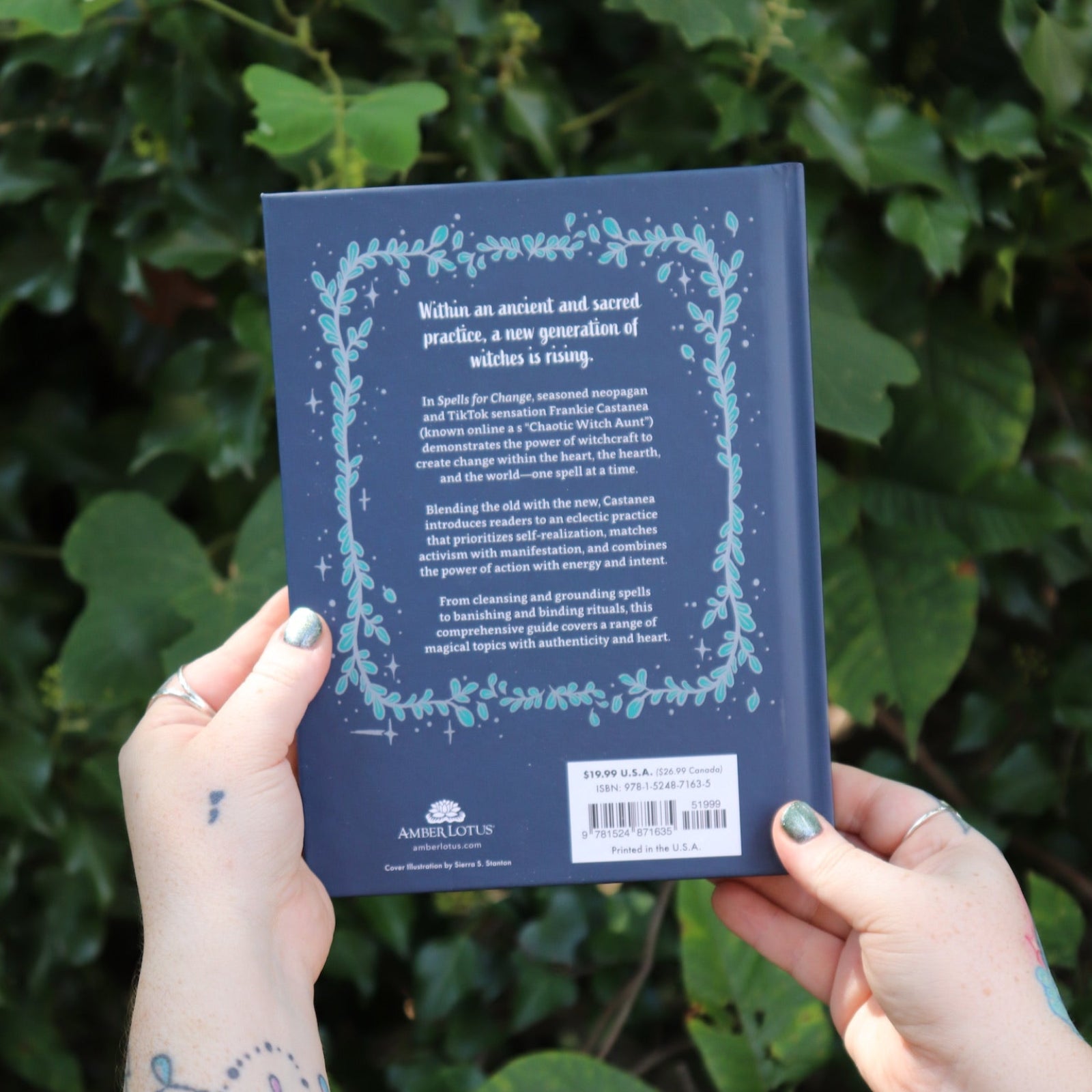 Someone holding the Spells for Change Hardcover Book in front of greenery, showing off the back.