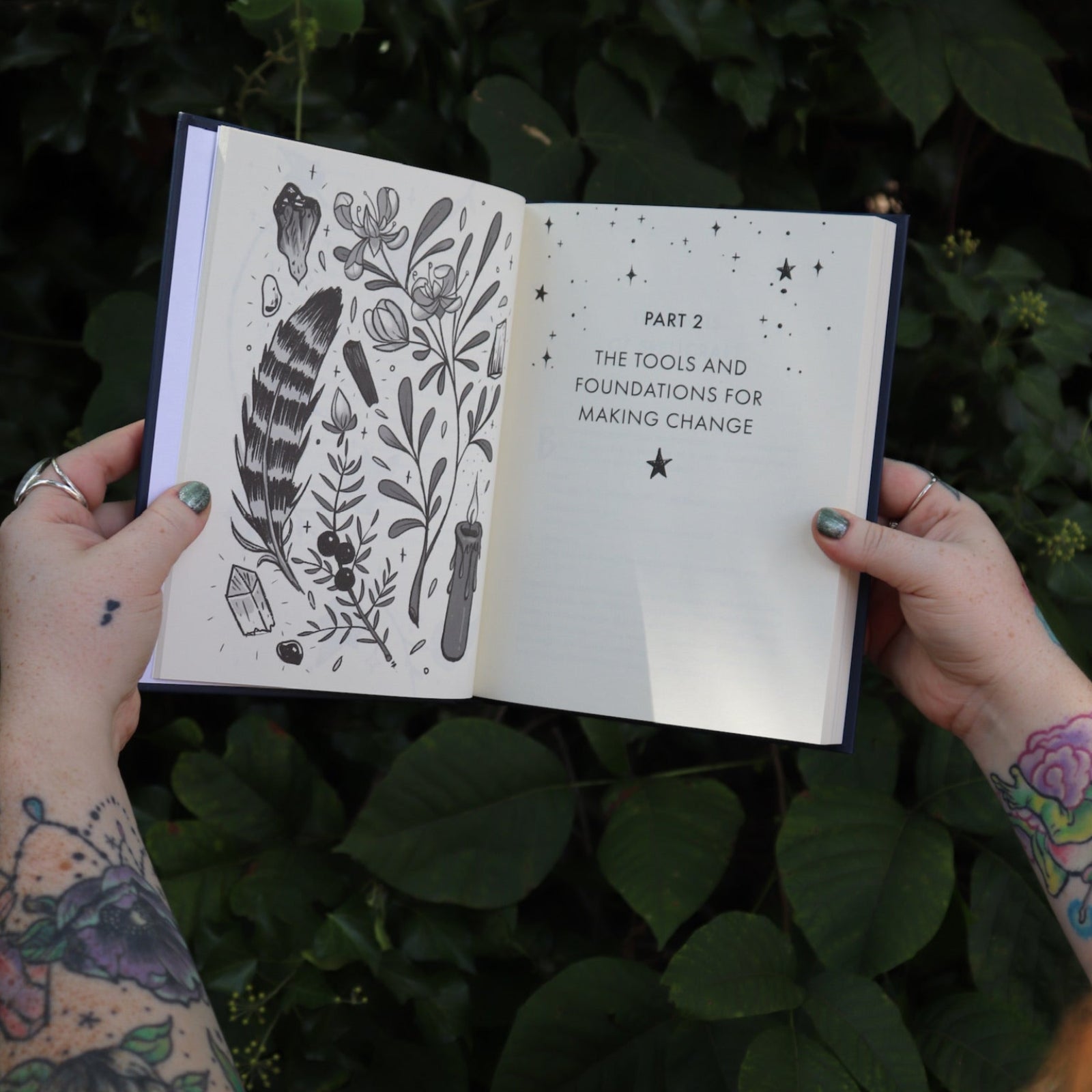 Someone holding the Spells for Change Hardcover Book open in front of greenery, showing off the pages.