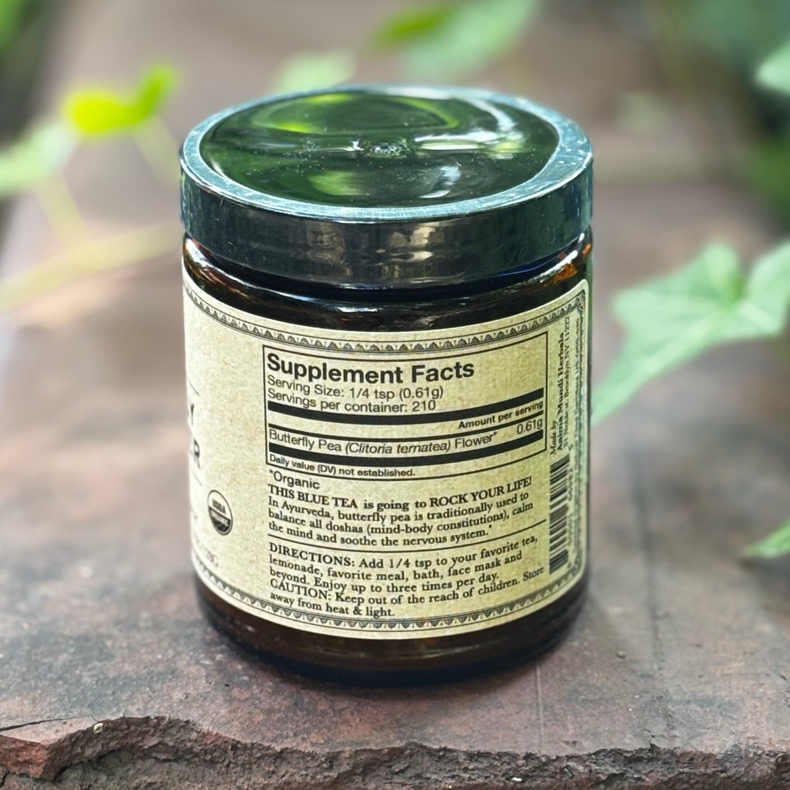 The back of the Blue Healer Butterfly Pea Flower Tea 4.5 oz Powder on a rusty red surface surrounded by green ivy.