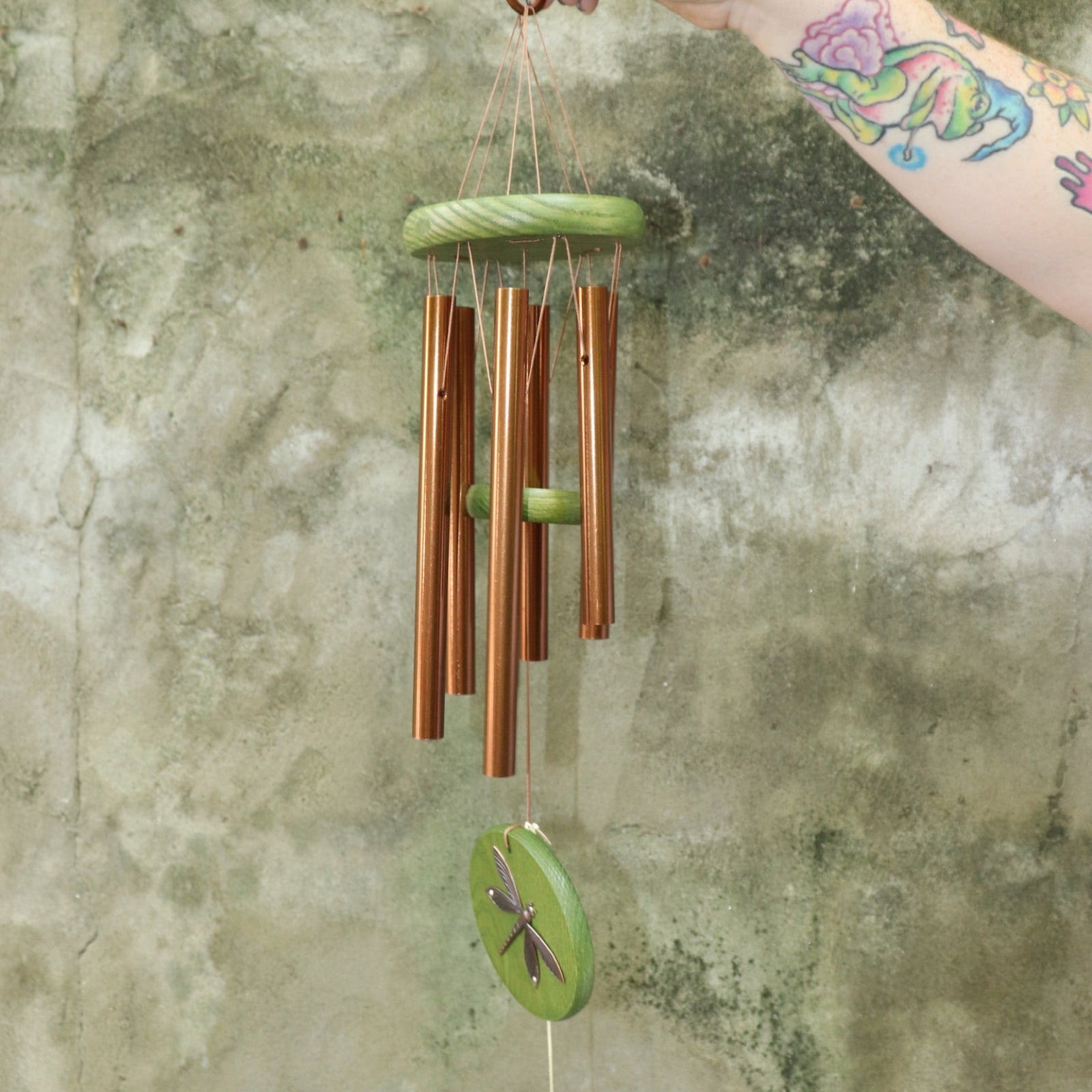 An up-close image of someone holding the Green Dragonfly Woodstock Habitats™ Wind Chime in front of a concrete wall.