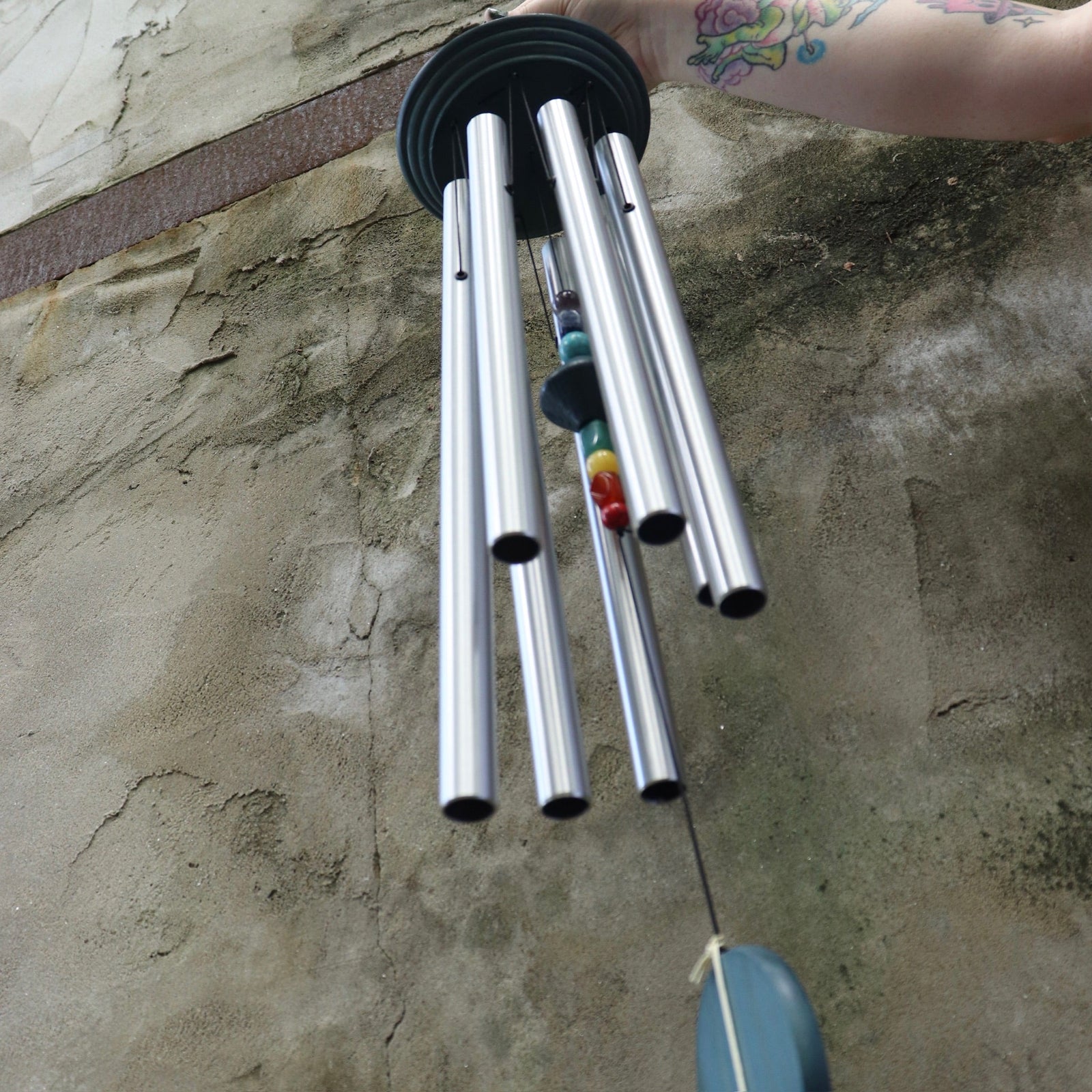 An up-close image of someone holding up the Seven Stone Blue Woodstock Chakra Chime in front of a concrete wall.