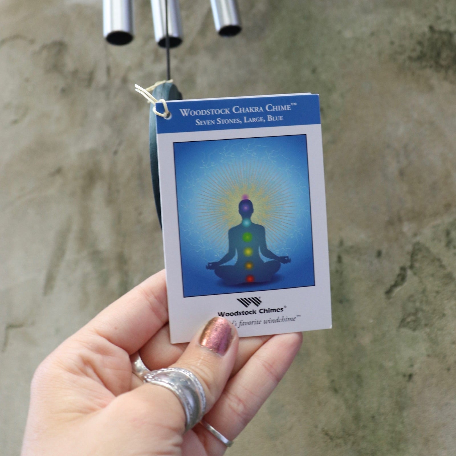 An up-close image of someone holding up the tag of the Seven Stone Blue Woodstock Chakra Chime in front of a concrete wall.