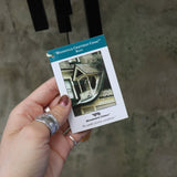 An up-close image of someone holding the tag of the Black Woodstock Craftsman Chime in front of a concrete wall.