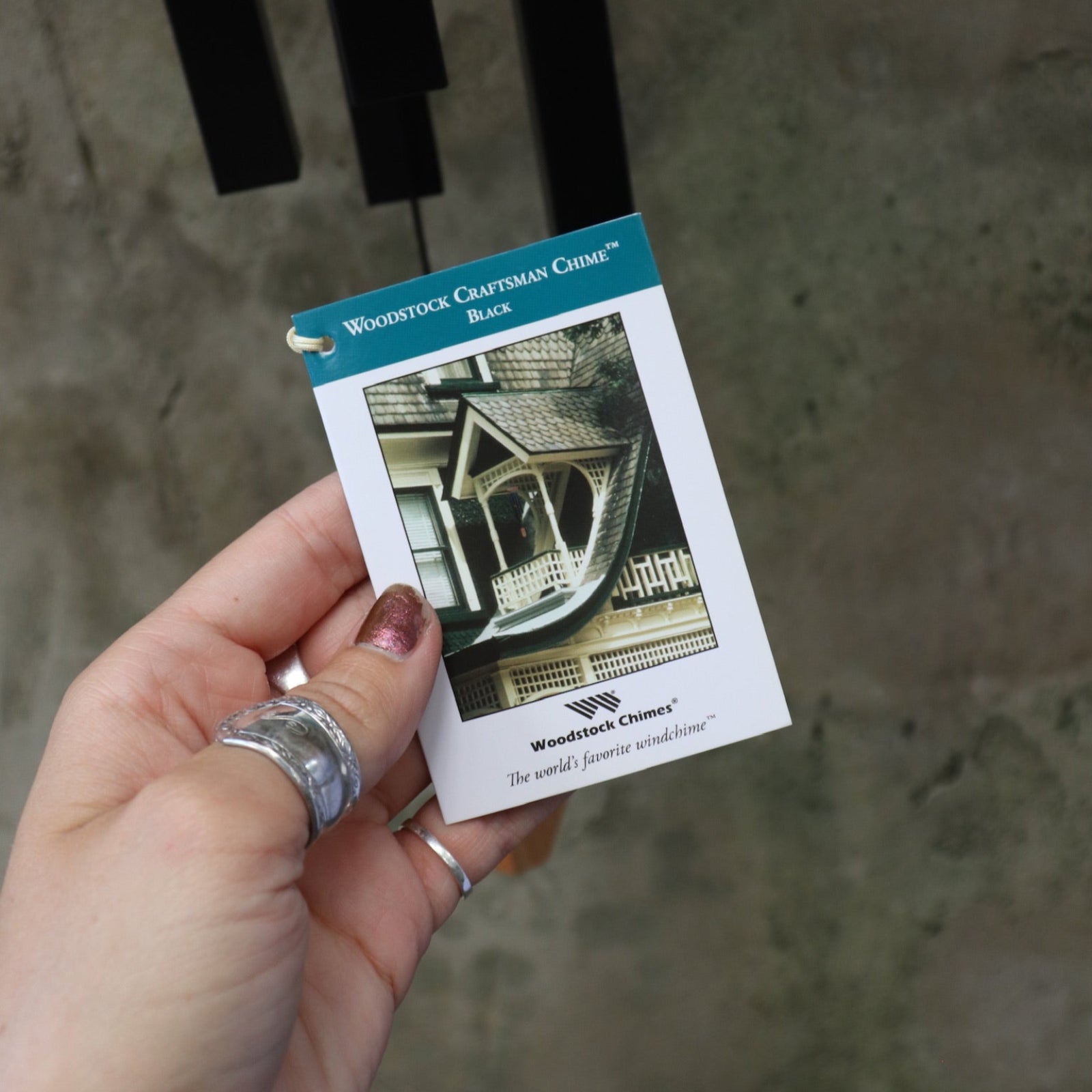 An up-close image of someone holding the tag of the Black Woodstock Craftsman Chime in front of a concrete wall.
