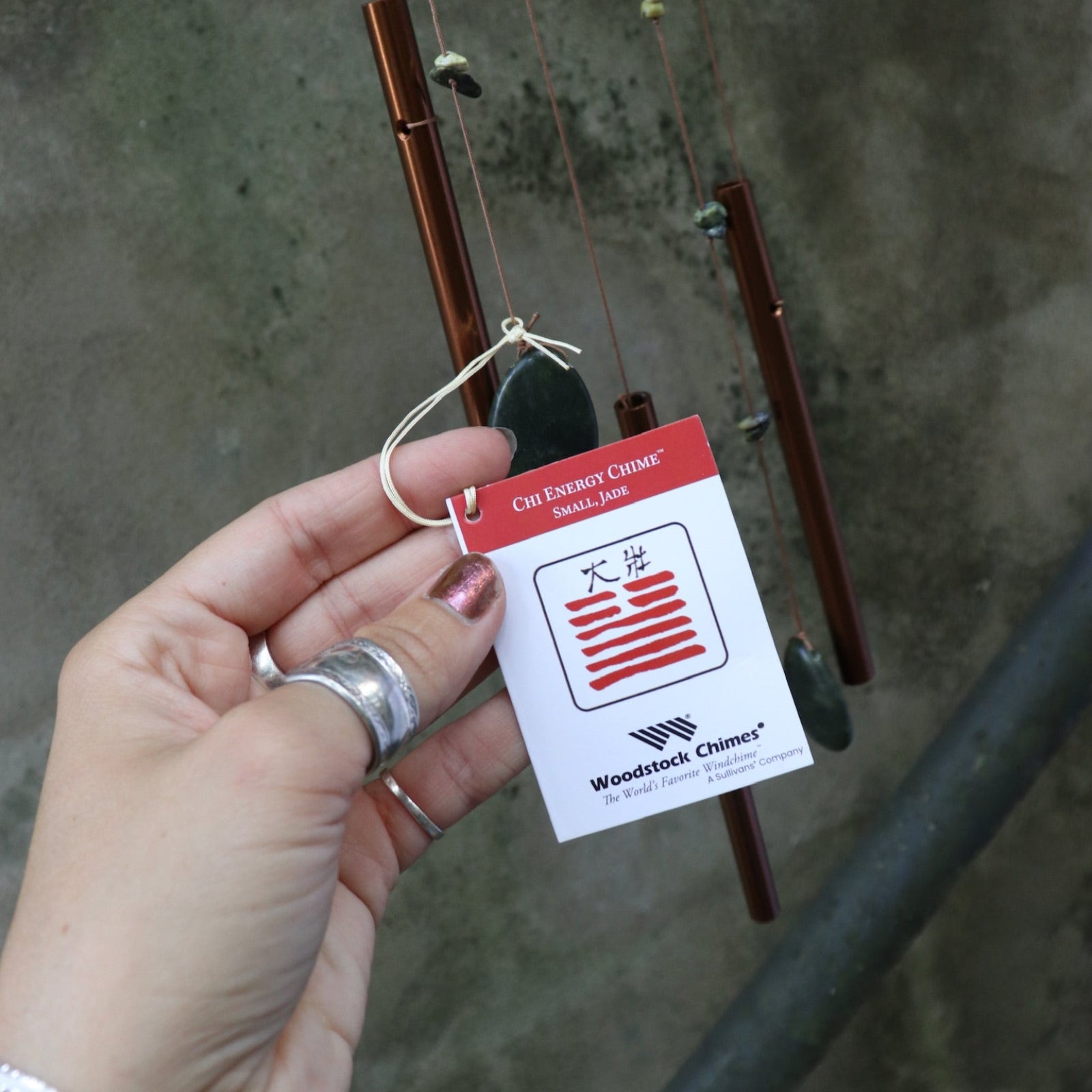 Someone holding the tag of the Small Jade Chi Energy Wind Chime in front of a concrete wall.