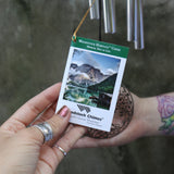 An up-close image of someone holding the tag of the Mirror Tree of Life Woodstock Habitats Wind Chime ornament in front of a cement wall.