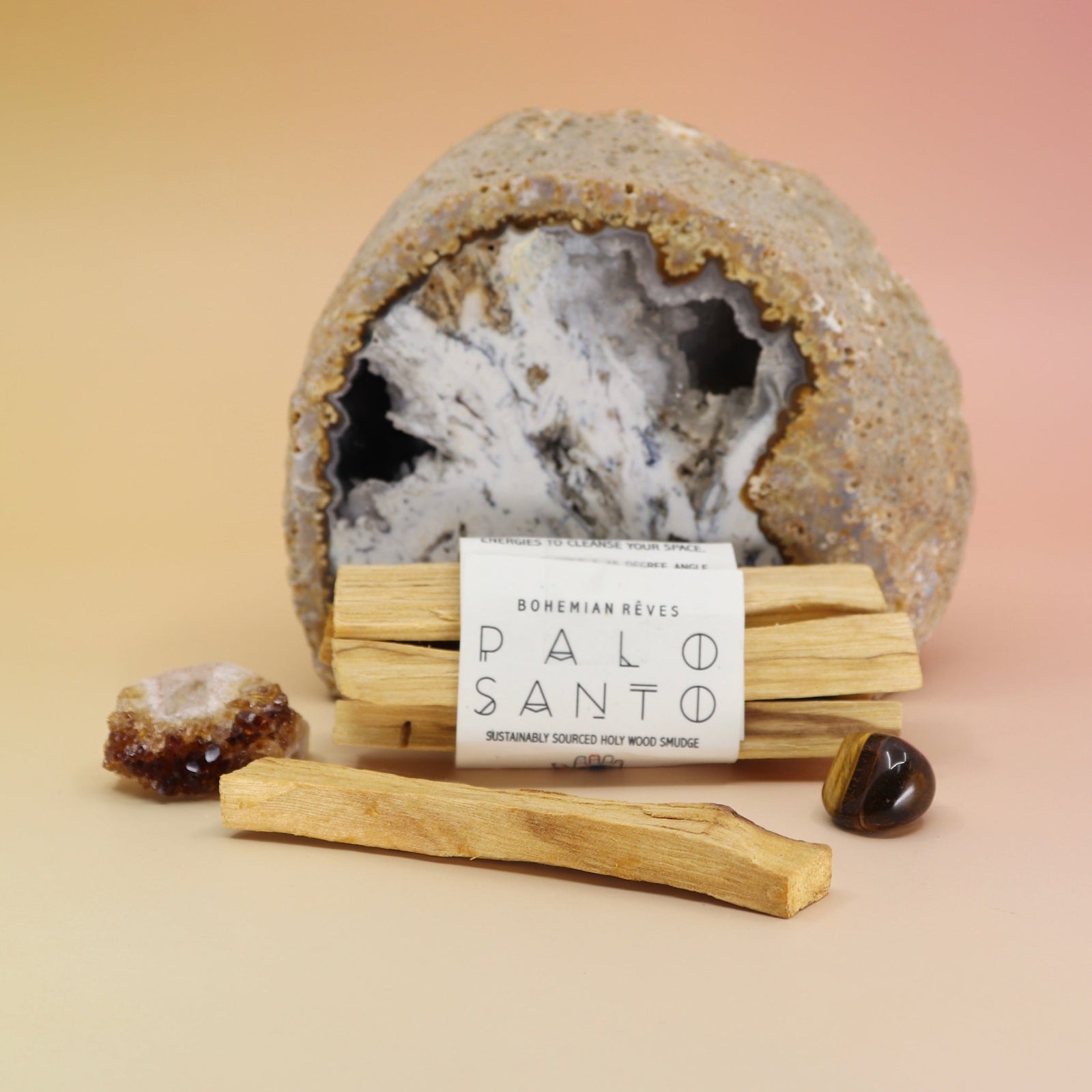 The Palo Santo Smudge Bundle by Bohemian Rêves in front of a cracked geode and surrounded by crystals all on a pink and yellow background.