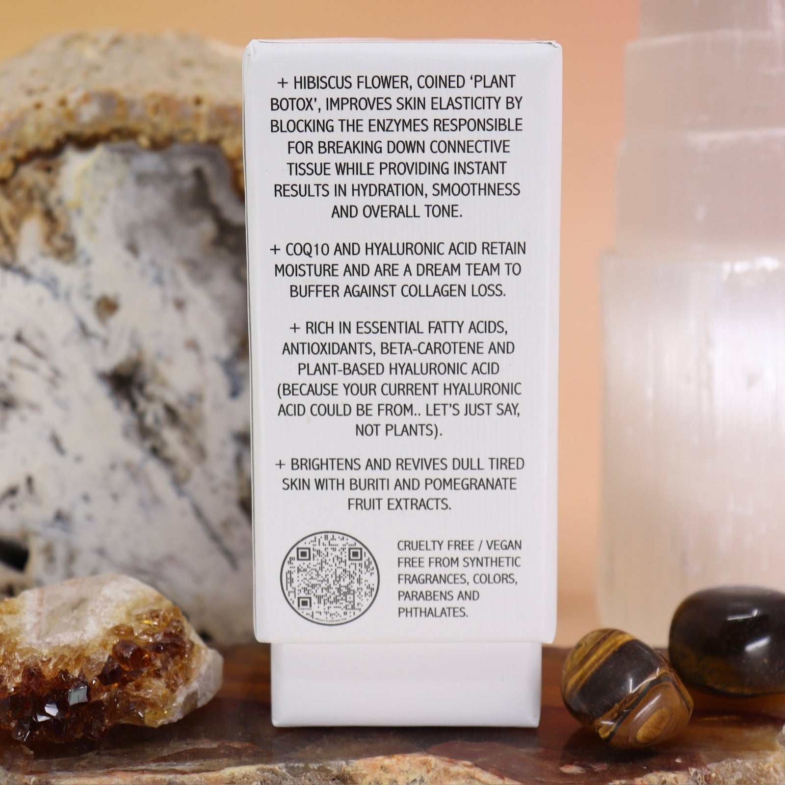 The side of the Anti-Aging Elixir Face Oil from Bohemian Rêves on a slice of agate in front of a open geode and selenite tower, all on a pink and yellow background.