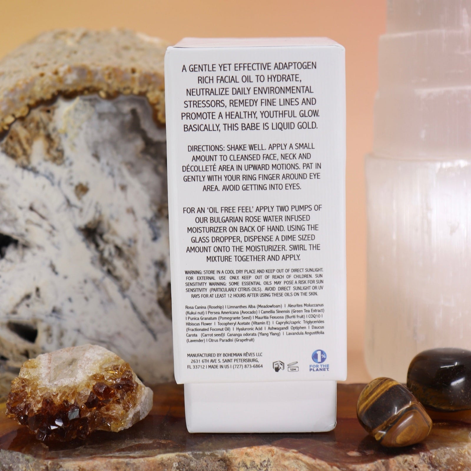 The back of the Anti-Aging Elixir Face Oil from Bohemian Rêves on a slice of agate in front of a open geode and selenite tower, all on a pink and yellow background.