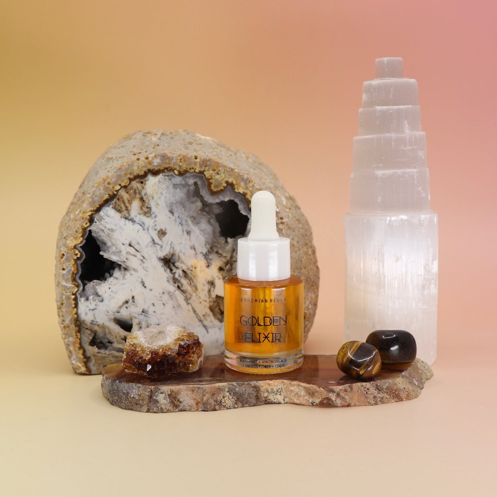 The Anti-Aging Elixir Face Oil from Bohemian Rêves out of its box and on a slice of agate in front of a open geode and selenite tower, all on a pink and yellow background.