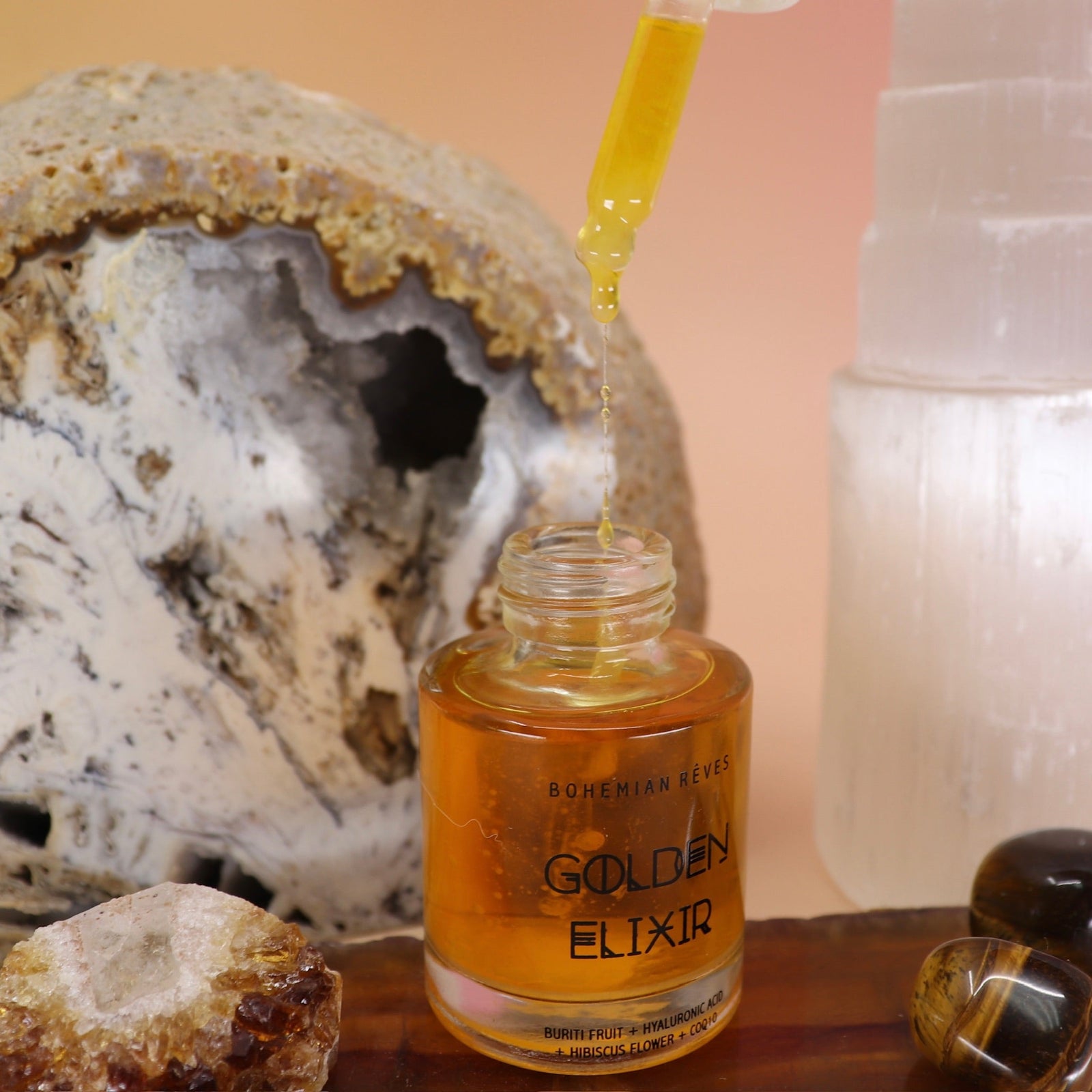 The Anti-Aging Elixir Face Oil from Bohemian Rêves out of its box and on a slice of agate in front of a open geode and selenite tower, all on a pink and yellow background.