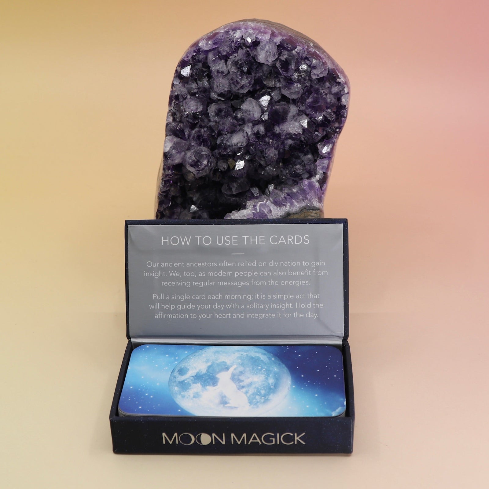 The Moon Magick Affirmation + Divination Cards against an amethyst piece all on a pink and yellow background.